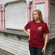 Lataa kuva Galleria-katseluun, Tyttö jolla päällä viininpunainen t-paita jossa pieni hirvipainatus talon edustalla
