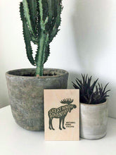 Lataa kuva Galleria-katseluun, puinen vaneripostikortti jossa metsänvihreä national parks finland hirvipainatus edestä ja takaa takana kaksi ruukkua ja kasvia
